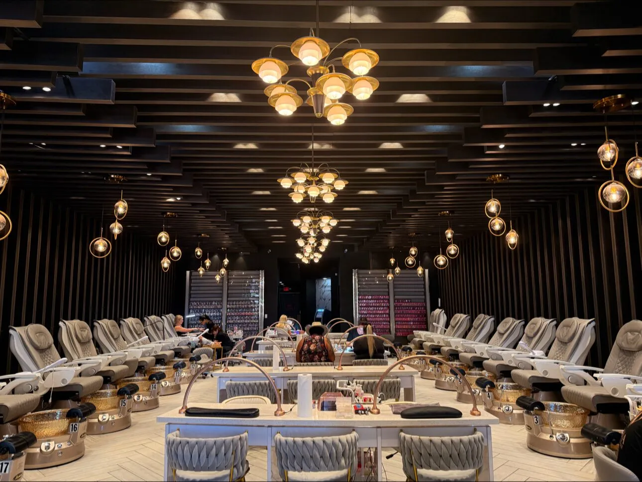 Cream pedicure chairs beneath gold chandeliers and a black slatted ceiling in the pedicure lounge at Aria Nail Bar Fashion Square.