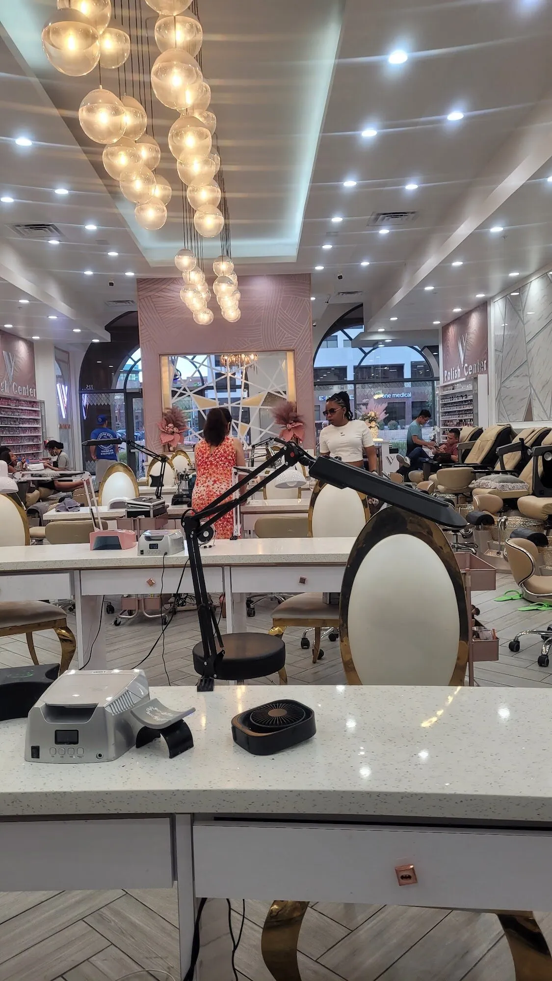 White manicure stations and gold-framed chairs beneath clusters of glass pendants in the manicure room at Aria Nail Bar Fashion Square.