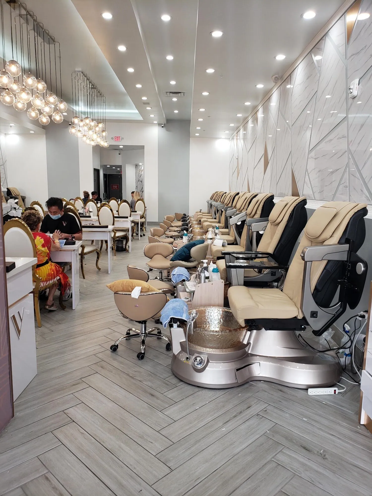 A row of cream massage pedicure chairs with gold basins along a white geometric wall at Aria Nail Bar Fashion Square.