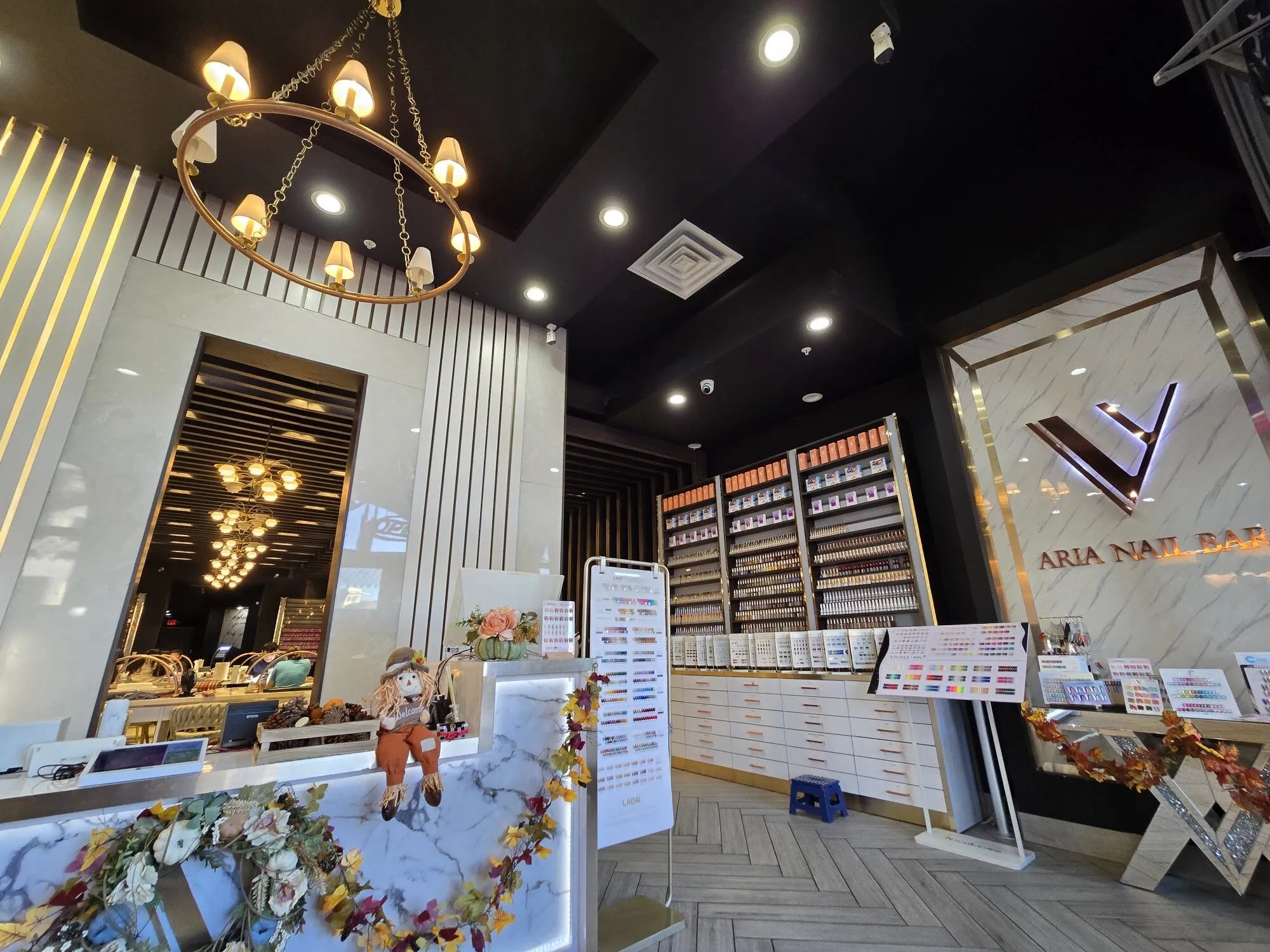 The reception at Aria Nail Bar Fashion Square: a marble counter beneath a gold ring chandelier, a wall of polish and the backlit Aria mark.