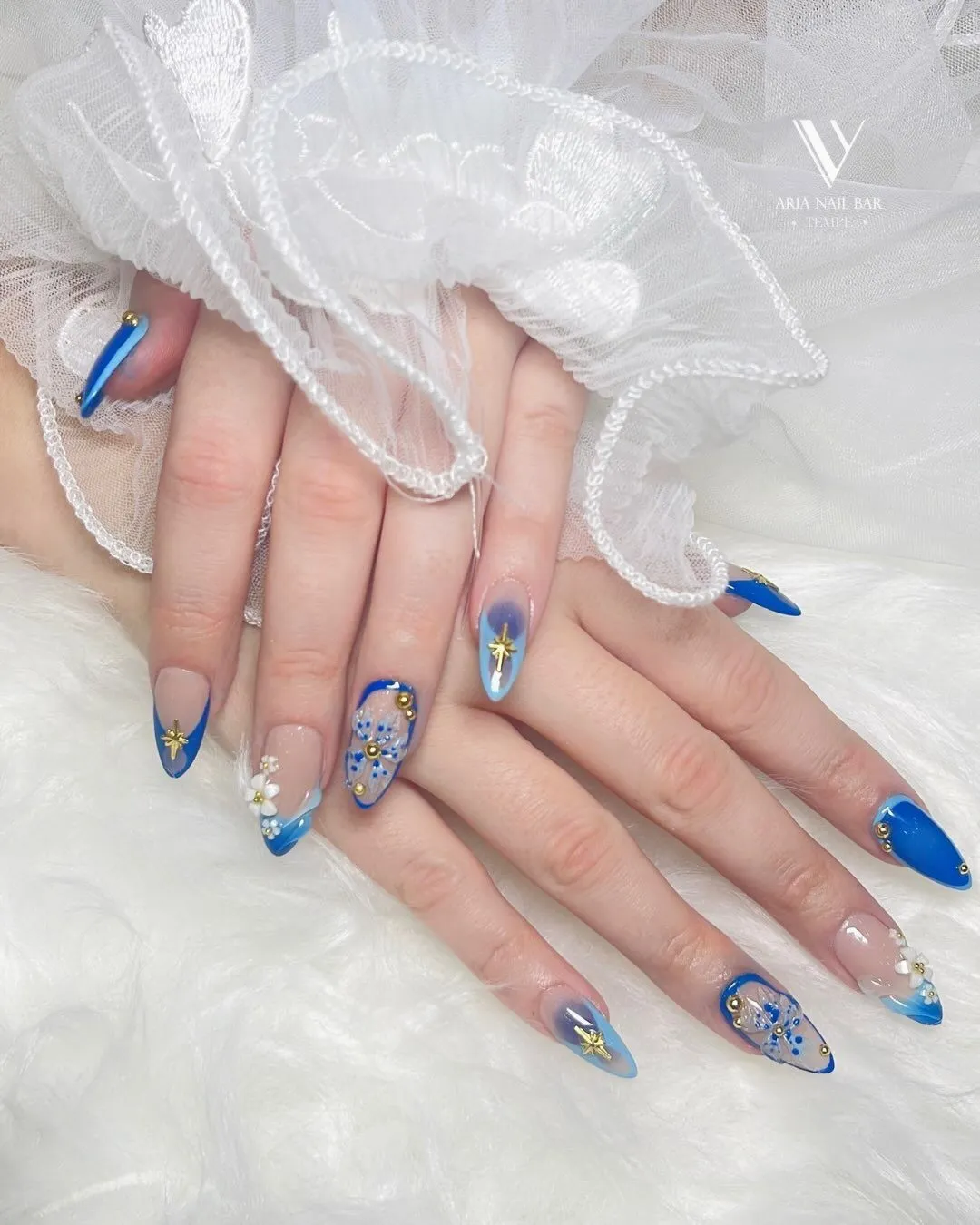 Blue aura French tips with hand-painted white flowers and gold stars on almond nails, resting on white tulle.