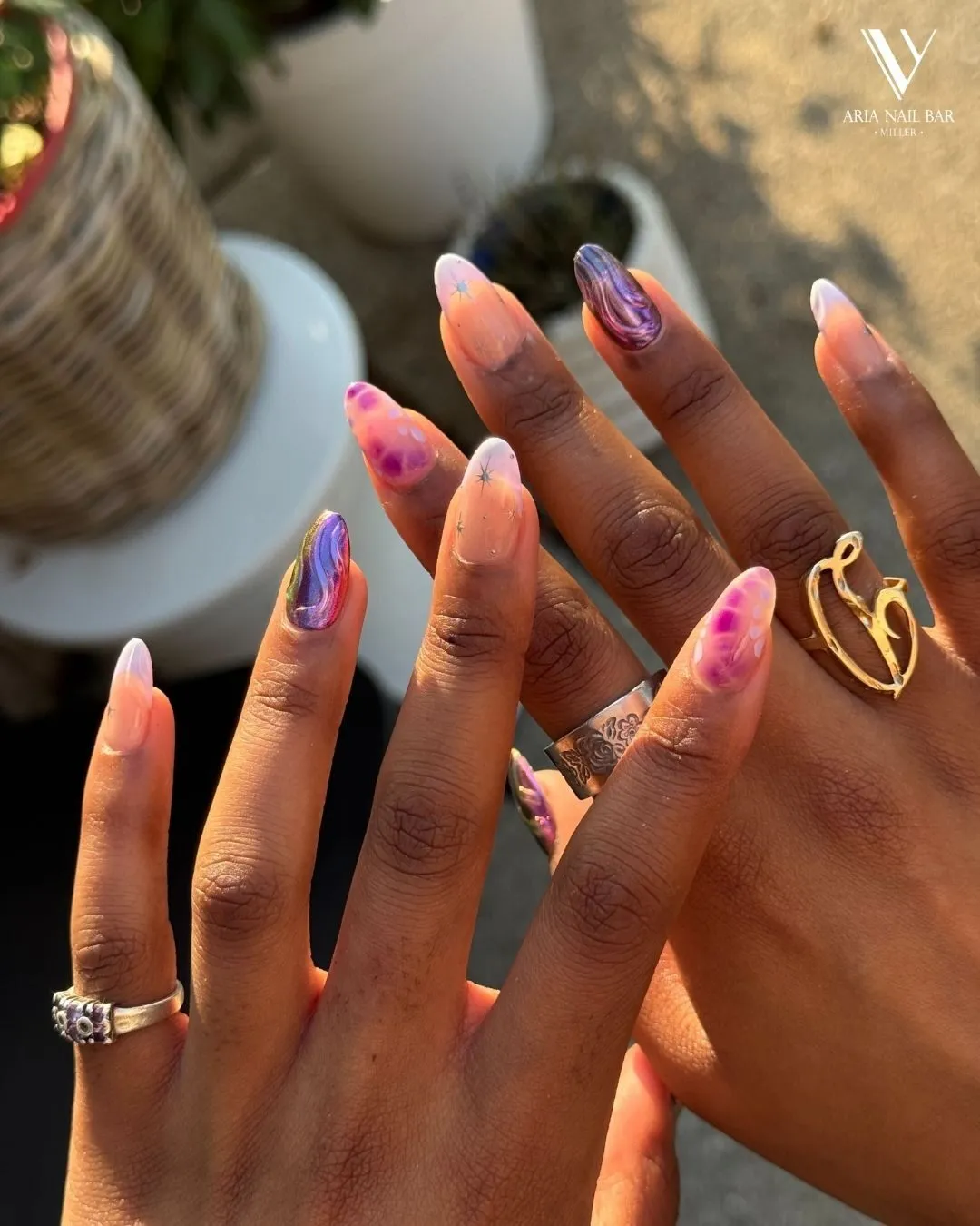 Long almond nails with purple and pink marbled aura art, styled with silver and gold rings.