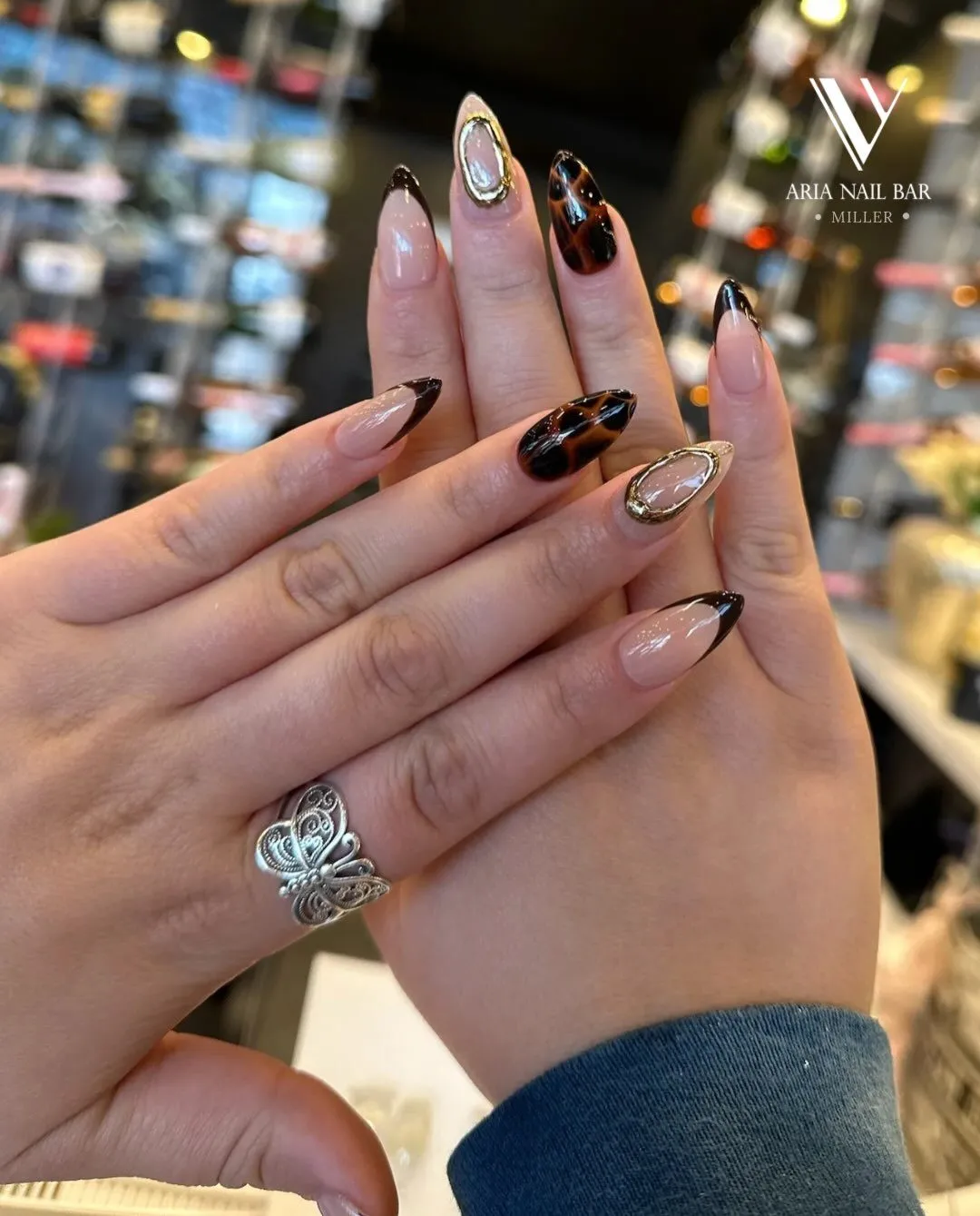 Short almond nails with black French tips and tortoiseshell accent nails framed in gold, in front of the polish wall.