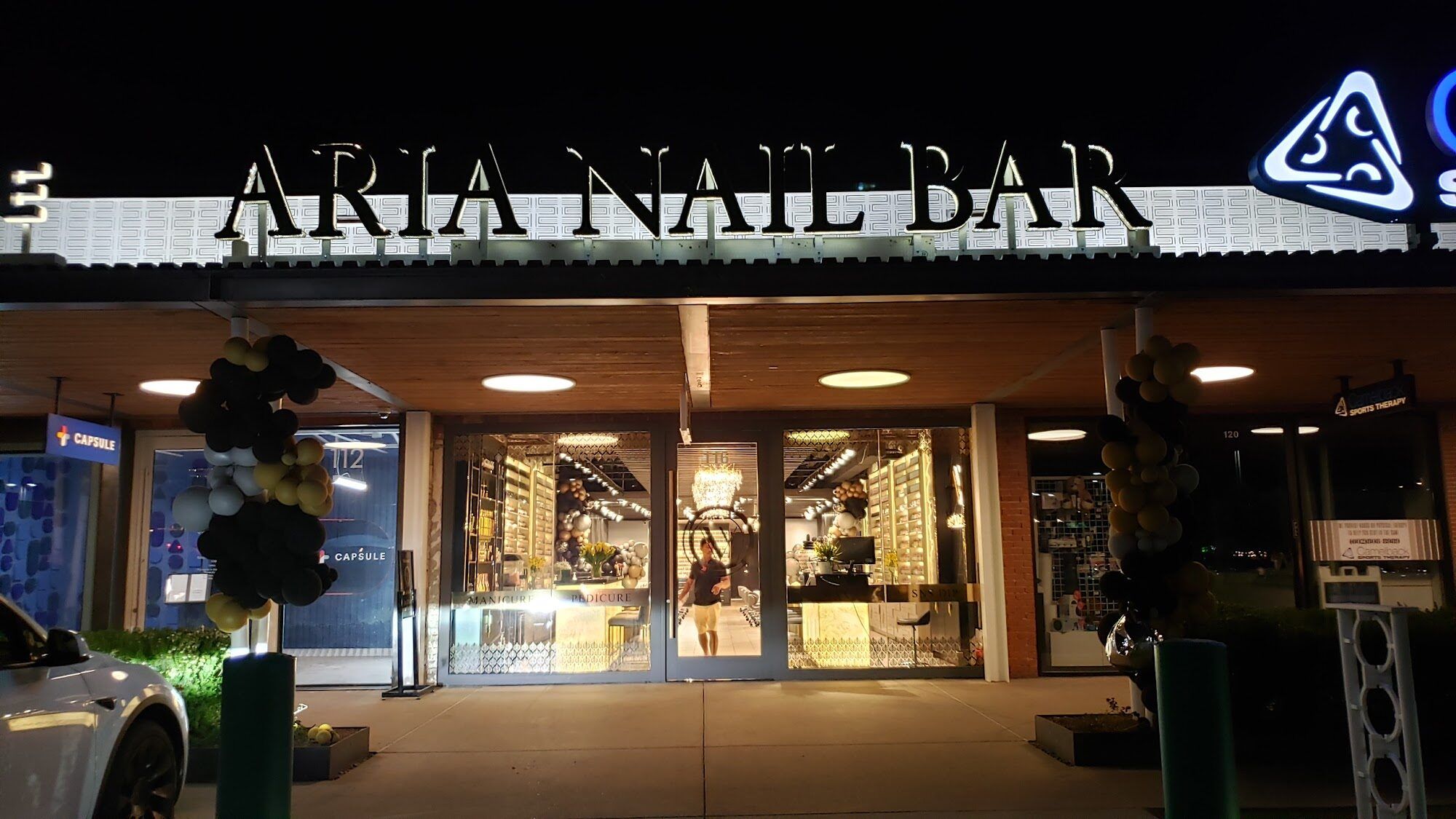 The storefront of Aria Nail Bar Phoenix Central at night, its illuminated sign above the glass entrance on Camelback Road.