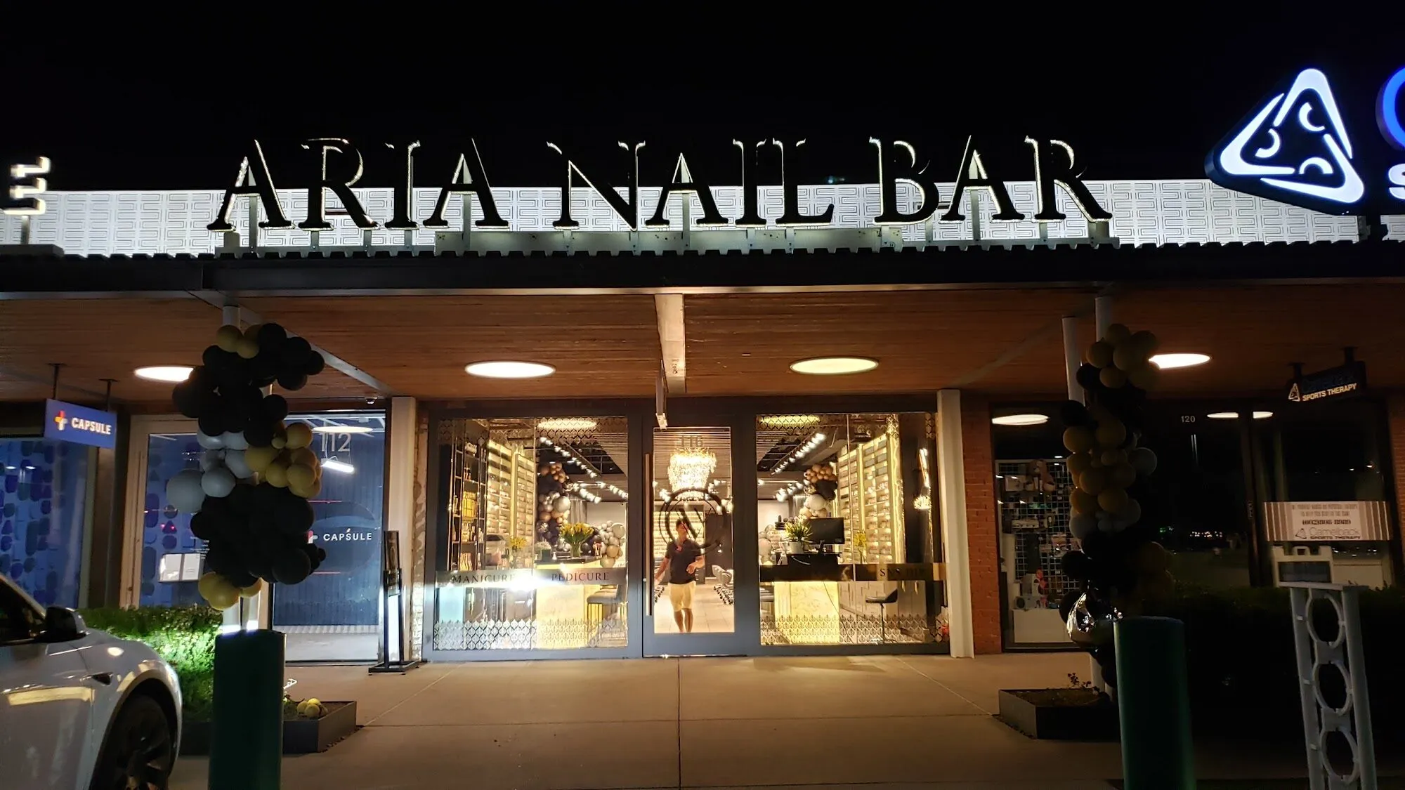 The storefront of Aria Nail Bar Phoenix Central at night, its illuminated sign above the glass entrance on Camelback Road.